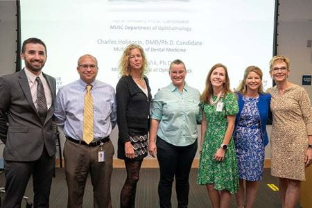  Group photo of people standing in front of a screen. They are Charles Holjencin, Andrew Jakymiw, Barb Rohrer, Kyrie Wilson, Jesse Goodwin, Lori McMahon and Lisa Saladin.