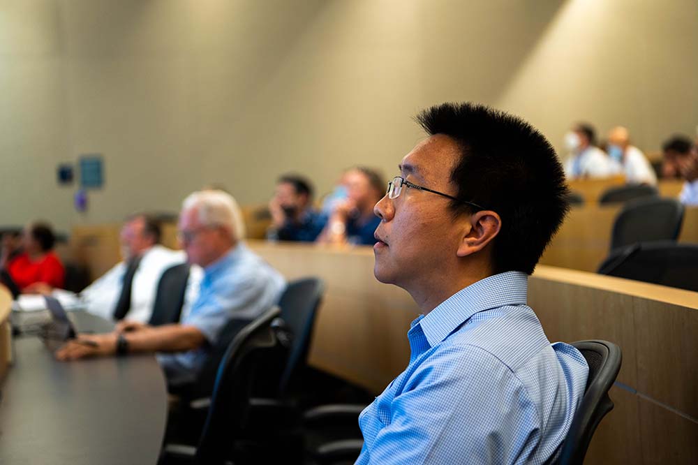 A man in a blud shirt and glasses listens intently in an auditorium.