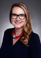 Smiling woman with long honey blonde hair, glasses and a black top poses for her headshot.