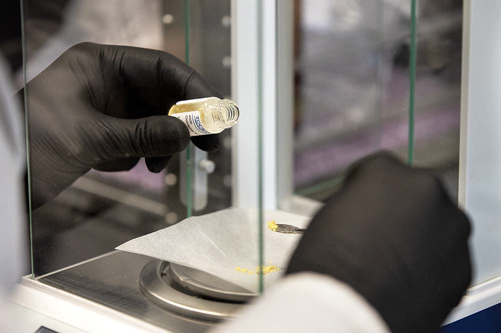 Hands wearing black lab gloves pour a vial of a chemical onto a scale.