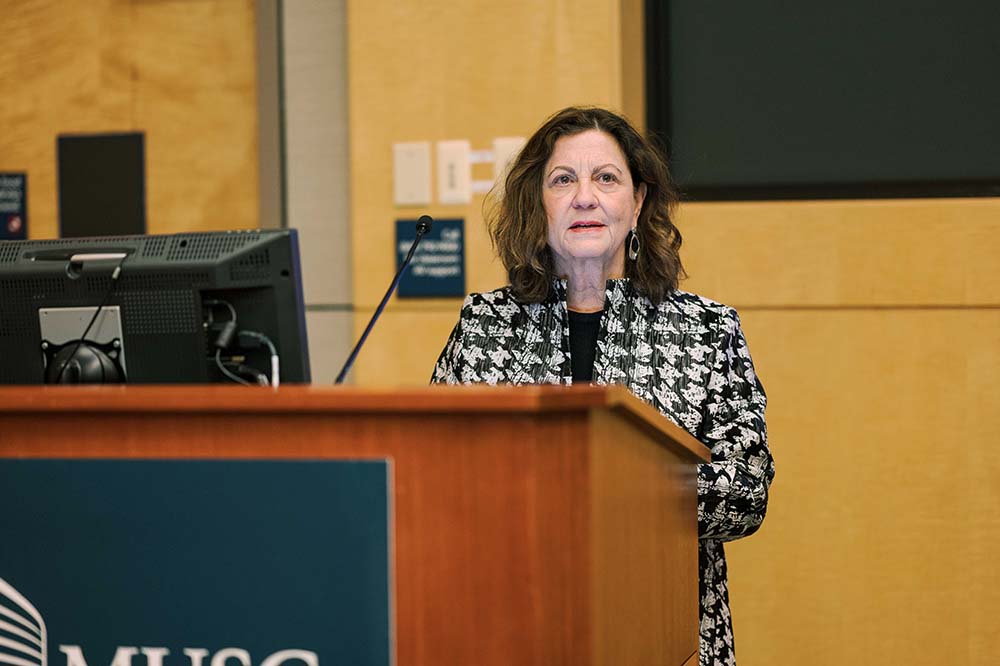 Woman wearing a checked jacket speaks at a podium.