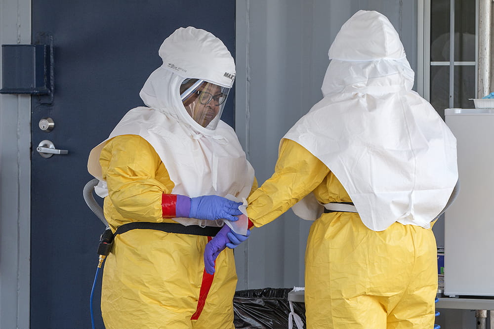 Two people fully covered by yellow jumpsuits and white head covers with clear masks. Both are wearing purple gloves.