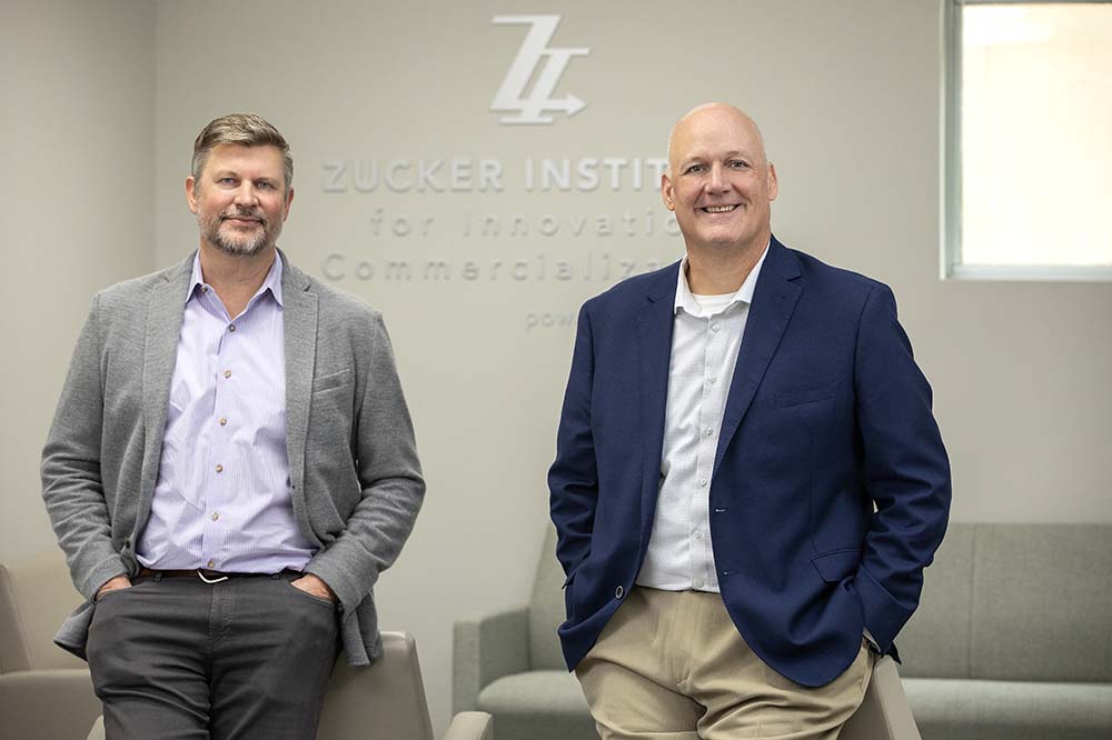 Two men wearing sport coats and dress shirts stand in front of a Zucker Institute sign.