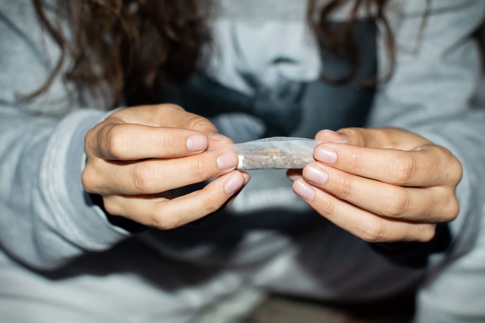 Young woman in tracksuit rolling a marijuana joint in the street at night. Details of hands rolling cannabis cigarette. Credit: José Antonio Luque Olmedo. Licensed from istockphoto.com