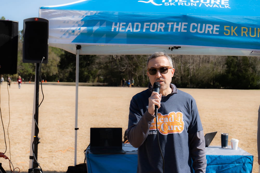 a man in a dark blue "Head for the Cure" shirt speaks into a microphone in front of a tent area set up in a large field