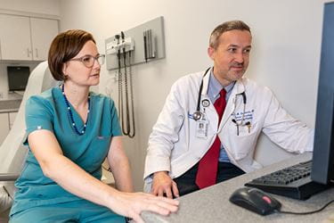 a male and female physician sit and look at a computer screen together in an exam area