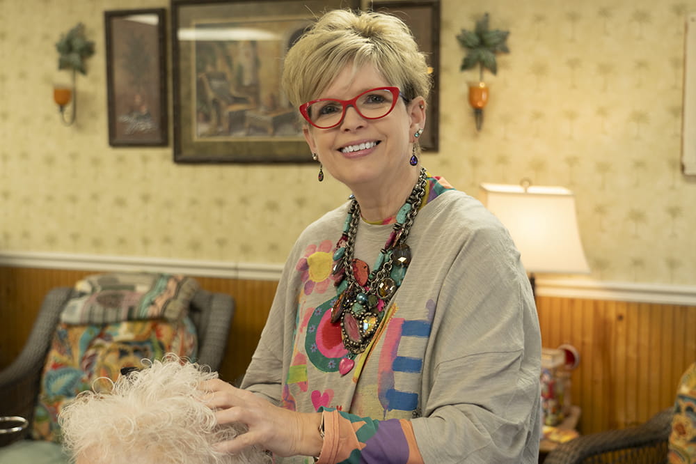 a woman with short blonde hair, red glasses and chunky necklaces pauses to smile at the camera over her client's head