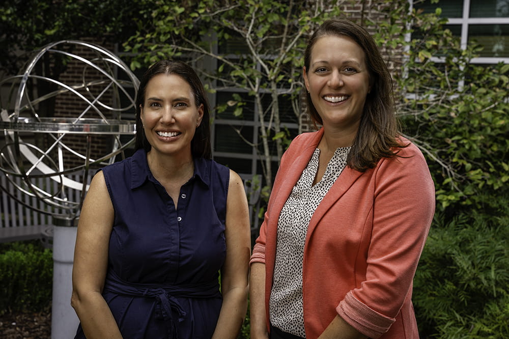 two women in business casual attire pose in a garden setting