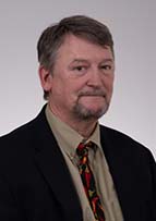 Headshot of man in dark suit and tan shirt. He has dark hear and a goatee.